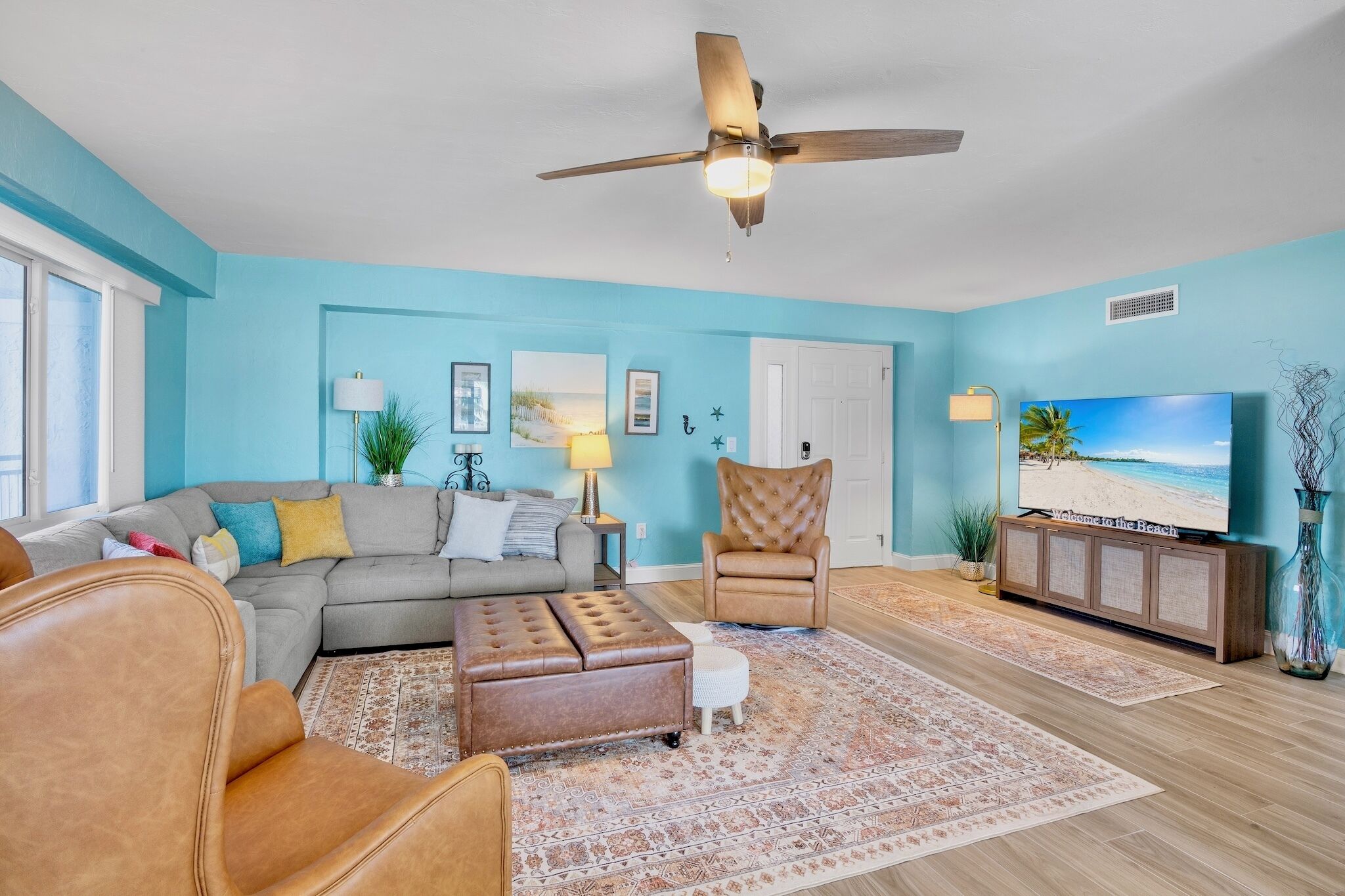 A living room with a sectional sofa, a leather armchair, a tufted ottoman, a television, and a ceiling fan.