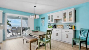 Een moderne keuken met witte kasten, een eethoek met een houten tafel en stoelen, en uitzicht op de oceaan door grote glazen deuren.