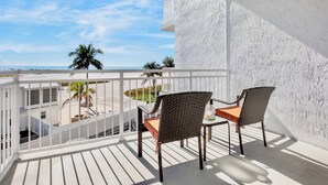 Een balkon met twee rieten stoelen en een tafel, met uitzicht op een strand met palmbomen en een heldere lucht.