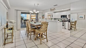 A modern kitchen with white cabinets, a central island, and a dining area with wooden chairs. - Beachfront Condo with Coastal Decor and Community Amenities - Jamaica Royale 306 (Siesta Key)