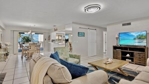 A spacious living area with a sofa, coffee table, and a TV. There is a dining area with chairs and a table, and a view of the outdoors through large windows. - Beachfront Condo with Coastal Decor and Community Amenities - Jamaica Royale 306 (Siesta Key)