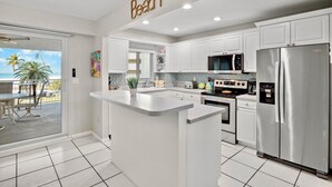 A modern kitchen with white cabinets, a central island, stainless steel appliances, and a view of the beach through a sliding glass door. - Beachfront Condo with Coastal Decor and Community Amenities - Jamaica Royale 306 (Siesta Key)