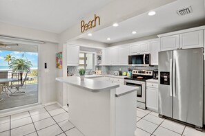 A modern kitchen with white cabinets, a central island, stainless steel appliances, and a view of the beach through a sliding glass door.