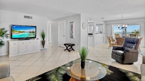 A modern living room with a flat-screen TV, a glass coffee table, a leather sofa, and a dining area with wicker furniture.