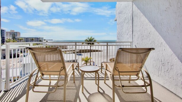 A balcony with two wicker chairs and a small table, overlooking a beach and cityscape.