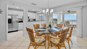 A modern kitchen with white cabinets and stainless steel appliances, a dining area with wicker chairs and a glass table, and a balcony with a view of the ocean.