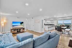 A modern living room with a large flat-screen TV, a sofa, and a dining area with a view of the ocean.