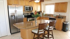 A kitchen with wooden cabinets, stainless steel appliances, a granite countertop, and two wicker chairs. - Jamaica Royale 502 Beautiful updates and great views! (Siesta Key)