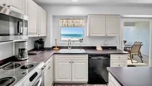 Een moderne keuken met witte kasten, een roestvrijstalen fornuis en een vaatwasser. Er is een raam met uitzicht op het strand.