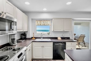 A modern kitchen with white cabinets, a stainless steel range, and a dishwasher. There's a window with a view of the beach.