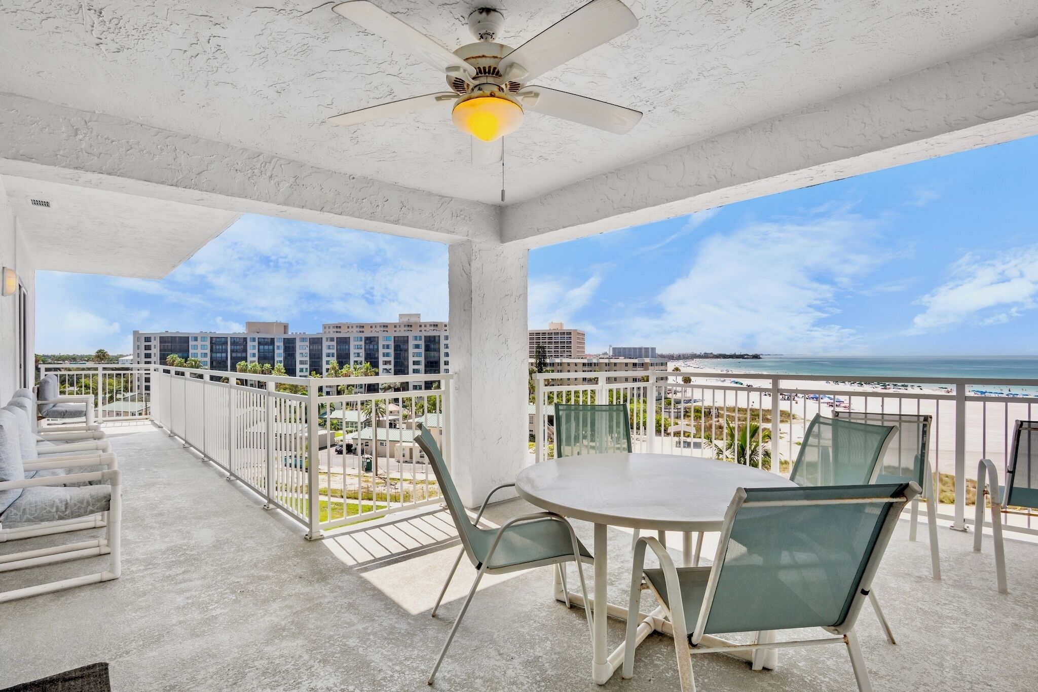 Un balcon avec une table et des chaises, un ventilateur de plafond et une vue sur la plage et les bâtiments.