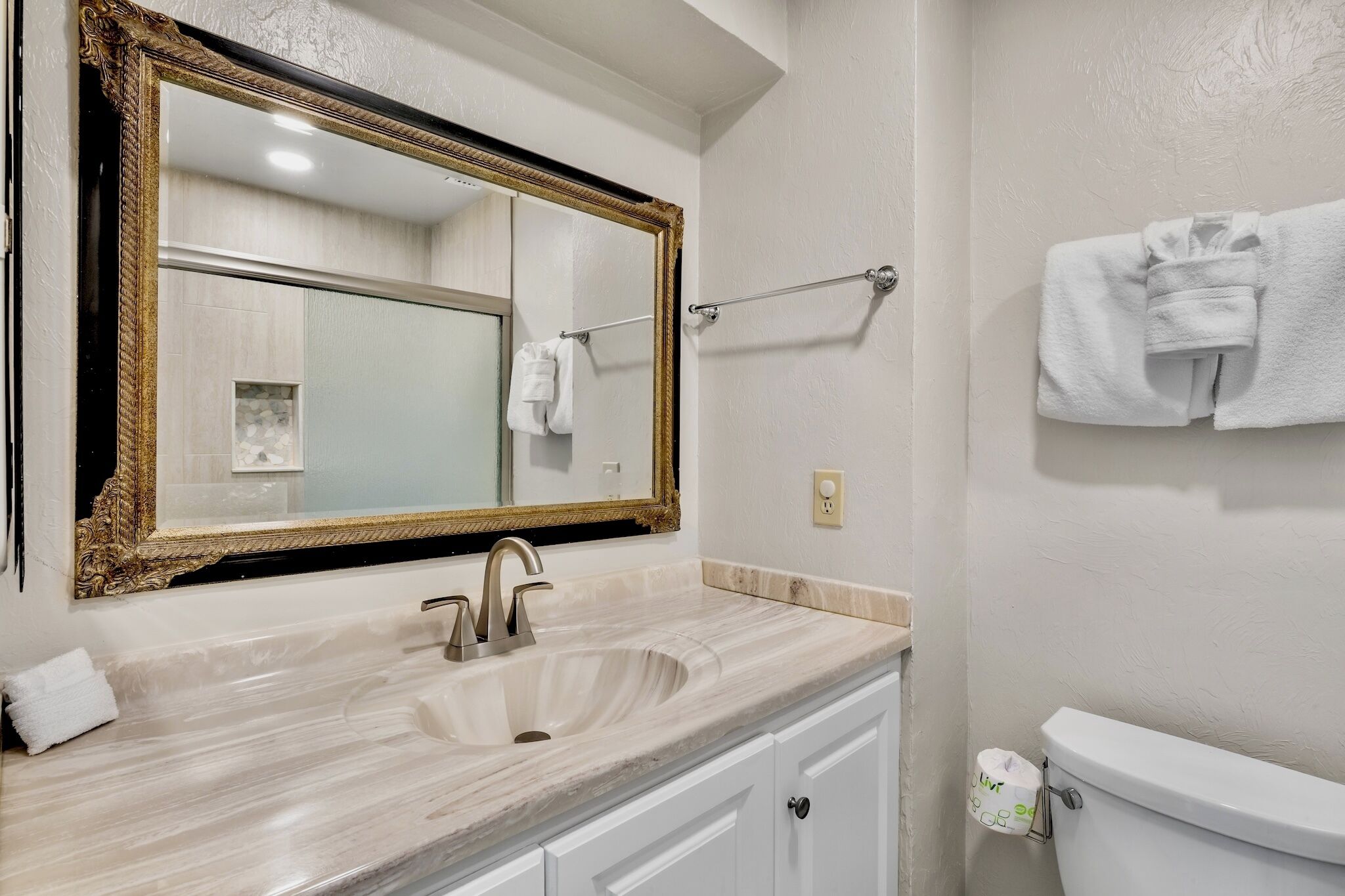 A bathroom with a marble sink, a gold-framed mirror, a towel rack, and a toilet with a toilet paper holder.