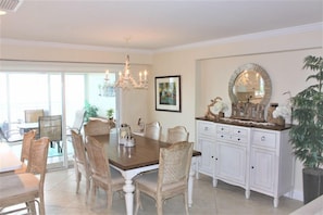 Un coin repas comprenant une table en bois, des chaises rembourrées, un lustre, un buffet blanc avec un miroir et une plante en pot.