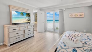 A bedroom with a TV, a bed with a patterned bedspread, a dresser, and a view of the ocean through a sliding door.