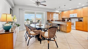 A modern kitchen with a dining area, large windows, and a ceiling fan.