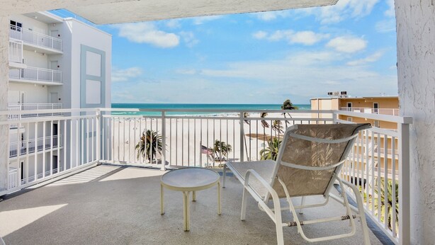 A balcony with a view of the beach, a white railing, a table, and a chair.
