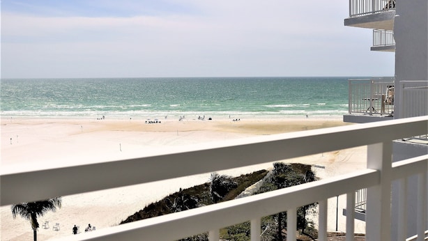 Vista a la playa desde un balcón con barandillas blancas, una playa de arena y el océano.