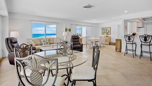 A spacious living area with a glass-top dining table, comfortable seating, and large windows offering a view of the ocean. - Couples Beachfront Getaway at Jamaica Royale 801 in Siesta Key! (Siesta Key)