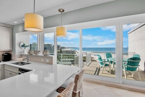 Une cuisine moderne dotée d’un grand îlot central, d’un coin repas avec des chaises vertes et d’un balcon offrant une vue sur la plage.