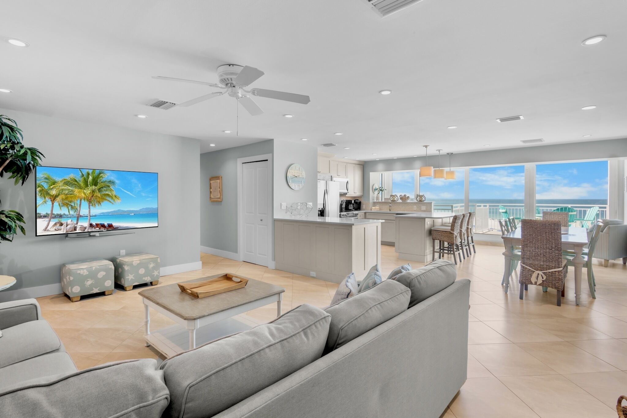 Un salon moderne comprenant un canapé, des poufs, une table basse, un téléviseur diffusant une scène de plage, un ventilateur de plafond et un coin repas avec des chaises et une table.