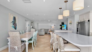 Cozinha moderna com ilha central, sala de jantar com cadeiras de vime e sala de estar com sofá e televisão.