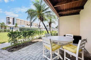 Un patio con una mesa redonda y sillas blancas, un área techada con techo de madera y vistas a palmeras y edificios al fondo.