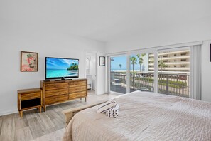 A bedroom with a bed, a TV, a wooden dresser, and a balcony with a view of palm trees and a building.