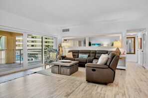 A spacious living room with a large sectional sofa, a coffee table, and a view of a pool and building through a glass door.