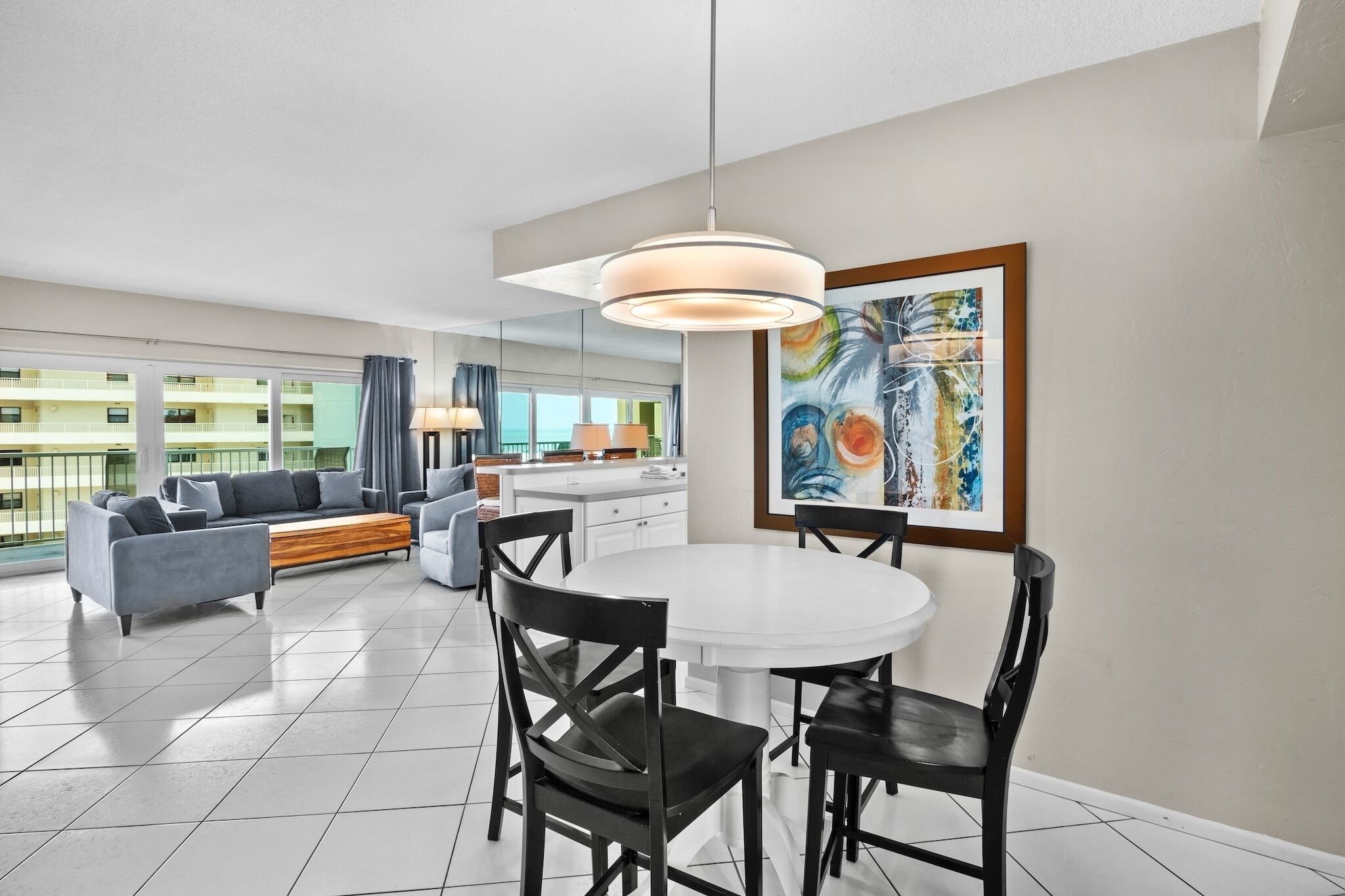 A modern living room with a dining area, featuring a white round table, black chairs, a gray sofa, and a large window with a view of a building.