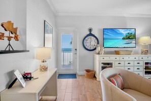 A living room with a sofa, a TV displaying a beach scene, a wall clock, and a door leading to a balcony.