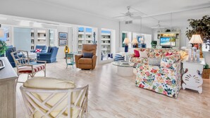 A spacious living room with a large window, a blue sofa, a floral couch, and a wooden chair.