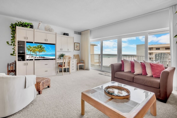 A modern living room with a flat-screen TV, a brown sofa with pink cushions, a glass coffee table, and a balcony with a view of the city.