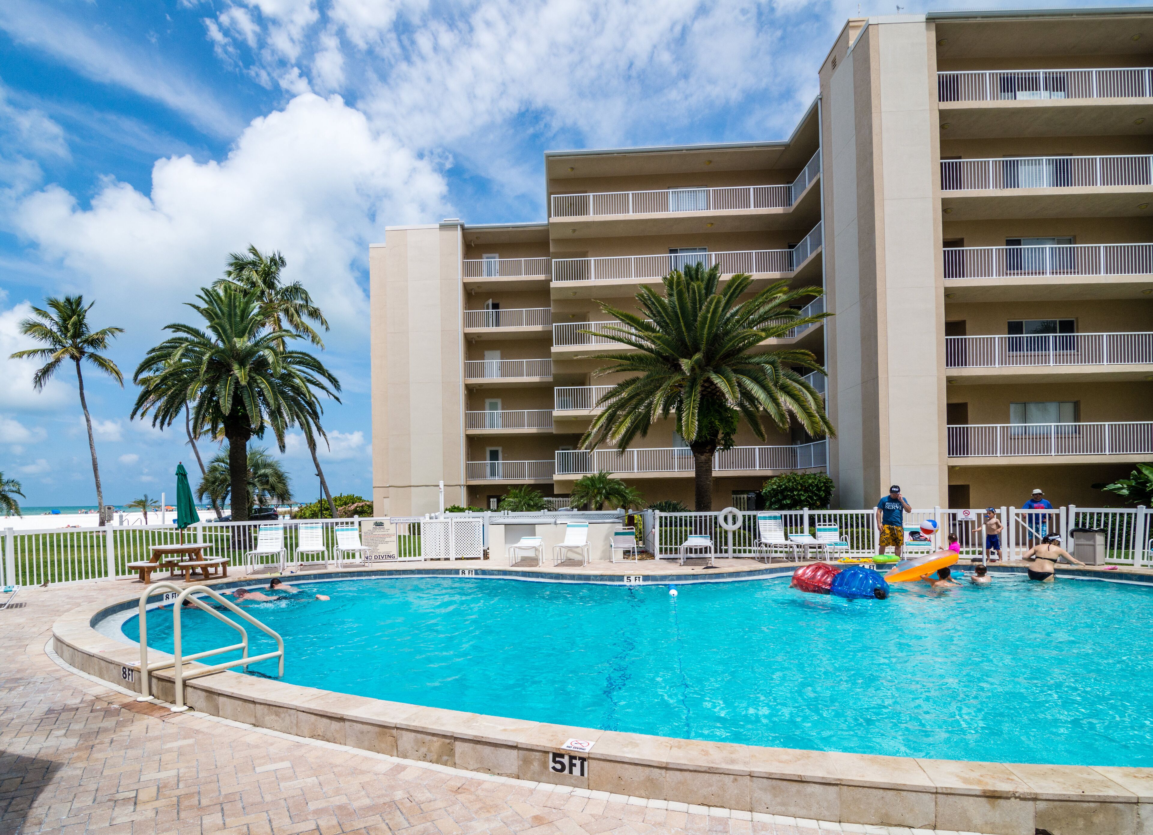 A swimming pool with a surrounding fence, palm trees, and a multi-story building with balconies.
