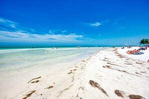 Une plage avec une eau turquoise et limpide, du sable blanc et quelques personnes visibles au loin.