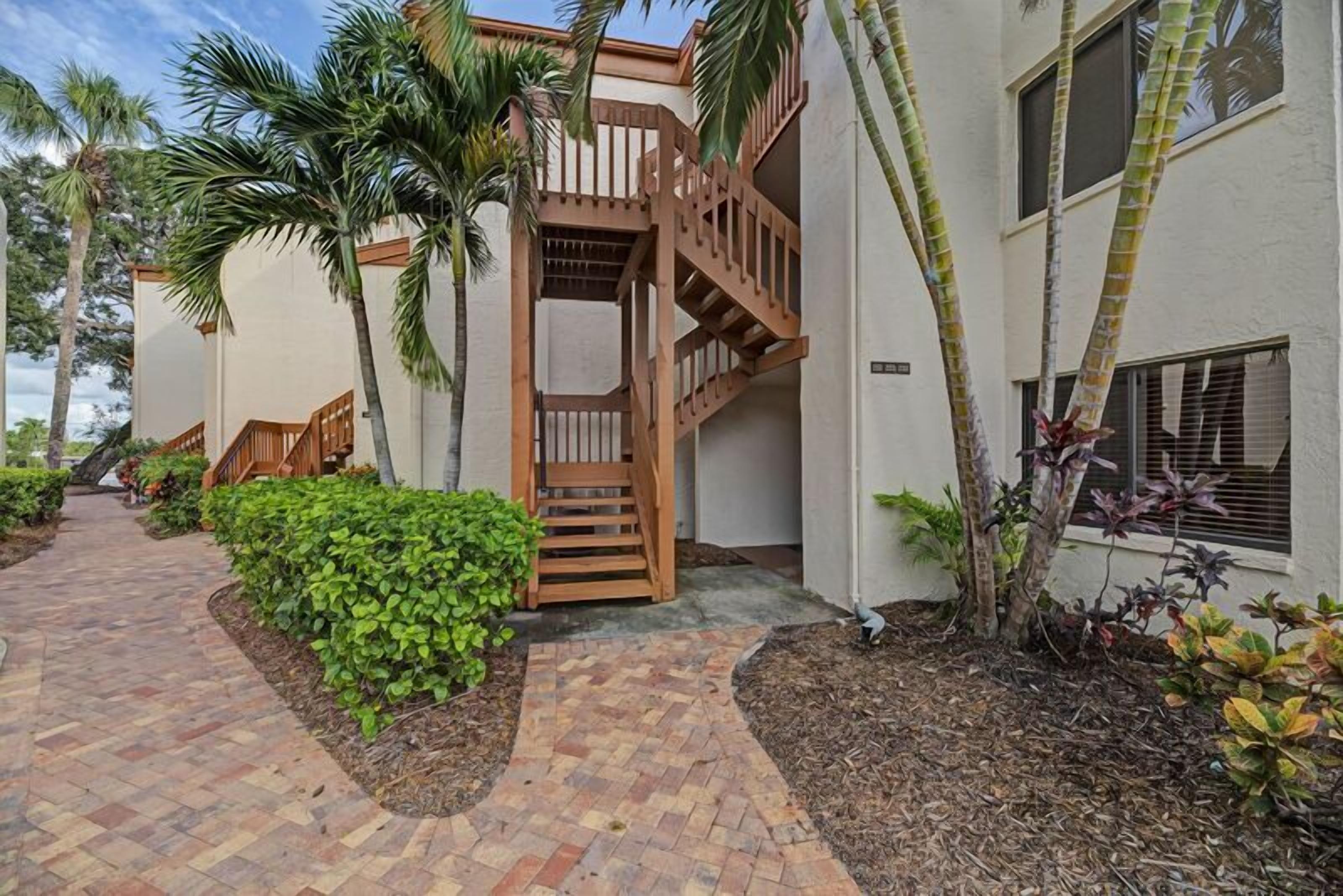 A building with a wooden staircase leading to a balcony, surrounded by palm trees and a brick pathway.