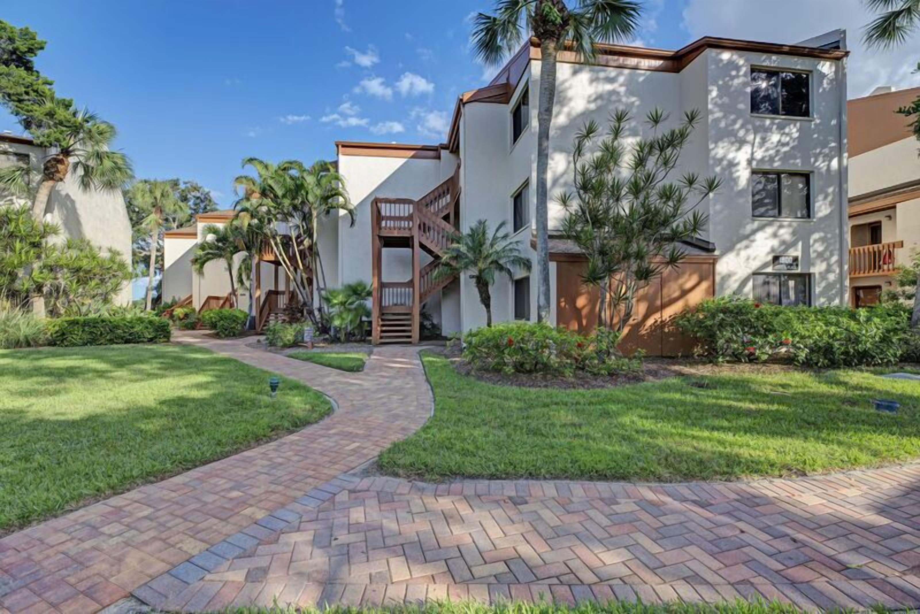 A modern building with a brick pathway leading to a wooden gate, surrounded by well-maintained grass and palm trees.
