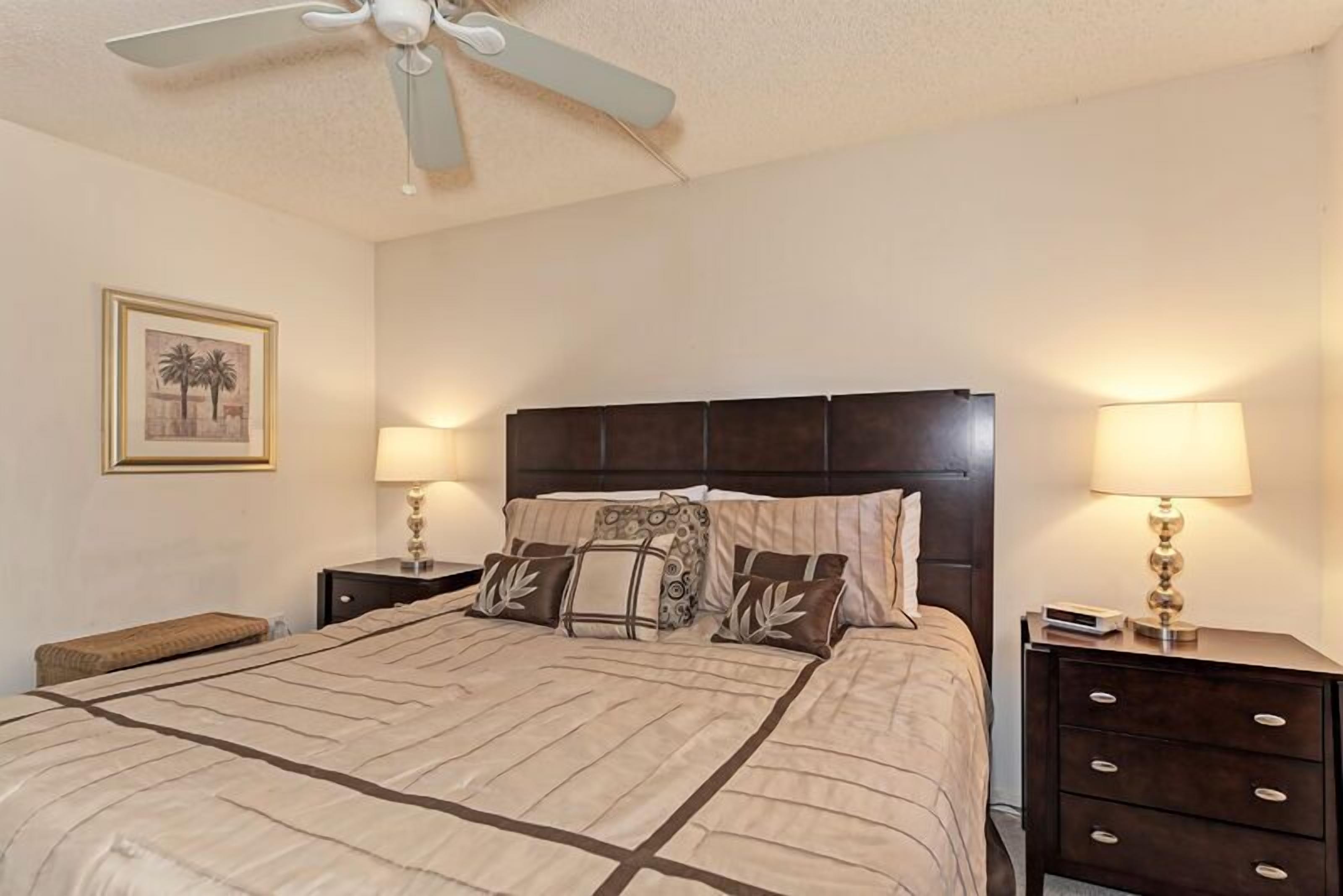 A neatly made bed with a dark headboard, two bedside tables with lamps, a ceiling fan, and a framed picture on the wall.