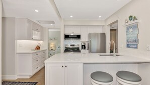 A modern kitchen with white cabinetry, a central island with bar stools, a stainless steel refrigerator, and a built-in oven. - Midnight Cove II 313F: Siesta Key Condo with Beach, Pool, Pickleball Access (Siesta Key)