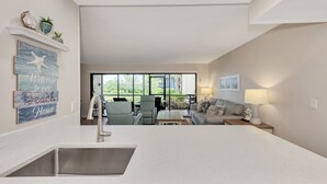 A modern kitchen with a large island, a dining area with a sofa and table, and a view of the outdoors through large windows. - Midnight Cove II 313F: Siesta Key Condo with Beach, Pool, Pickleball Access (Siesta Key)