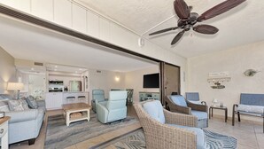 A spacious living area with a ceiling fan, light blue and wicker furniture, a kitchen in the background, and a flat-screen TV mounted on the wall. - Midnight Cove II 313F: Siesta Key Condo with Beach, Pool, Pickleball Access (Siesta Key)
