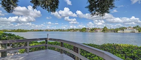A wooden deck with a railing overlooking a body of water, surrounded by greenery and buildings in the distance.