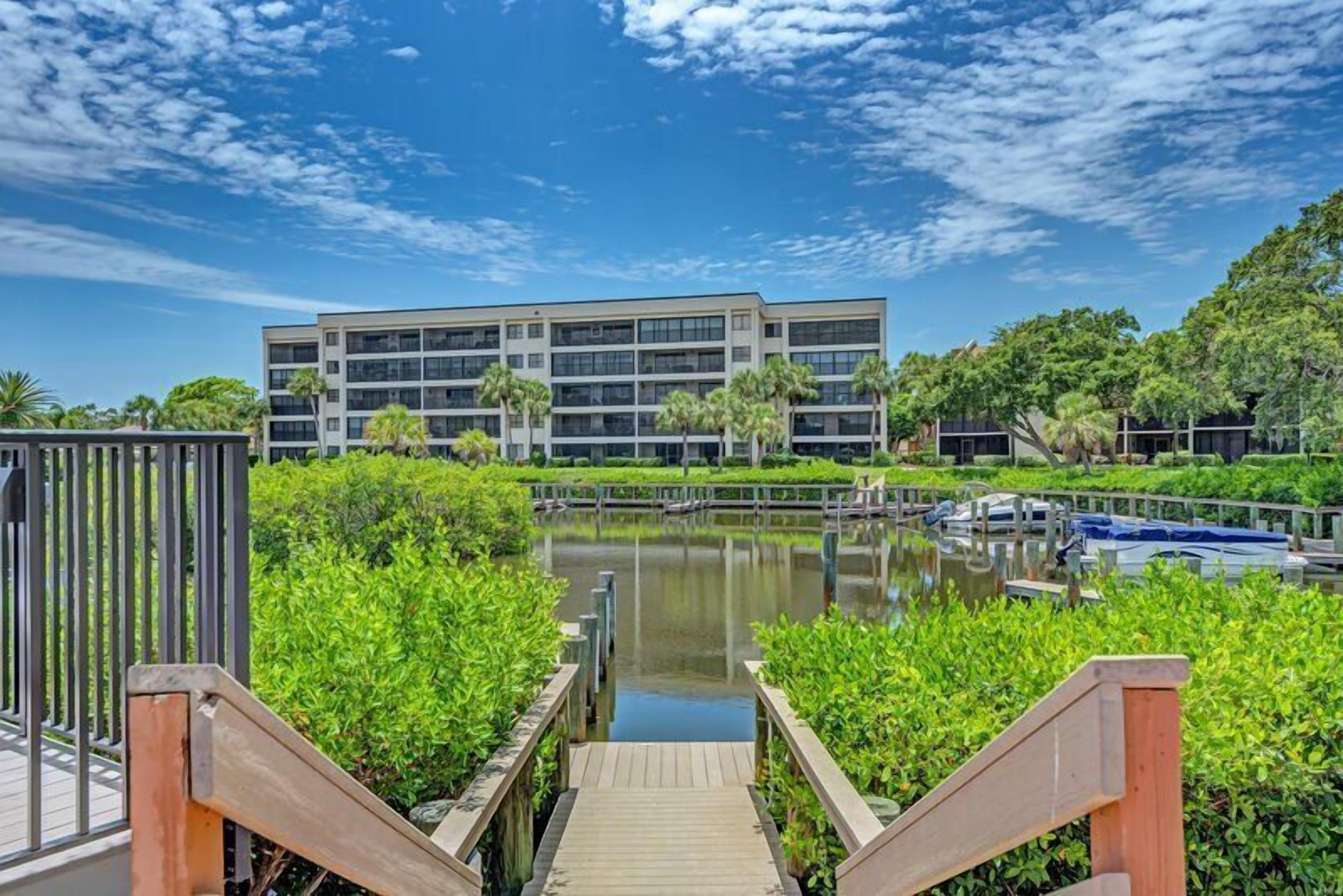A modern building with a water body in front, surrounded by greenery and a clear sky.