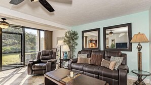 A spacious living room with a leather sofa, a recliner, and a coffee table. There are two ceiling fans, a large window, and a mirror on the wall. - Midnight Cove II 411F - Ground floor unit with Pool & Beach Access (Siesta Key)