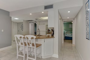 A compact kitchen with white cabinets, a built-in oven, and a sink. There are three white chairs with wooden seats. The kitchen leads to a bedroom with a bed and a wall with a sign.