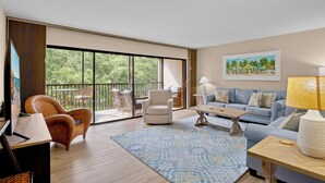Ein geräumiges Wohnzimmer mit einer großen Schiebeglastür, die auf einen Balkon im Freien führt, einem blauen Teppich mit Muster, einem hellblauen Sofa und einem Holztisch.