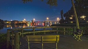 Eine Holzterrasse mit Bänken bietet Blick auf ruhiges Wasser bei Nacht, im Hintergrund sind Gebäude und Straßenlaternen zu sehen.