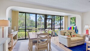 A spacious living area with a large window, a dining table, a sofa, and a TV. - Midnight Cove II 510F - Siesta Key Beach Condo Rental with Community Amenities (Siesta Key)