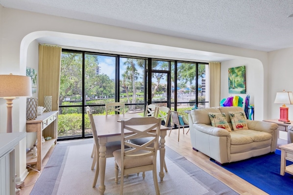 A spacious living area with a large window, a dining table, a sofa, and a TV. - Midnight Cove II 510F - Siesta Key Beach Condo Rental with Community Amenities (Siesta Key)