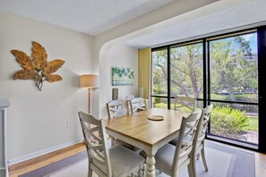 A dining area with a wooden table and chairs, a lamp, and a large window with a view of trees and buildings. - Midnight Cove II 510F - Siesta Key Beach Condo Rental with Community Amenities (Siesta Key)