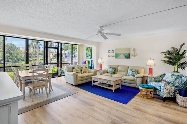 A spacious living room with a large window, a ceiling fan, and a dining area with a table and chairs. - Midnight Cove II 510F - Siesta Key Beach Condo Rental with Community Amenities (Siesta Key)